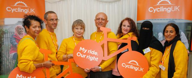 A group of people all wearing knitted yellow sweaters are standing together in front of Cycling UK branded boards and holding cardboard bikes with the words 'My Ride. Our Right' in one wheel and the Cycling UK logo in the other