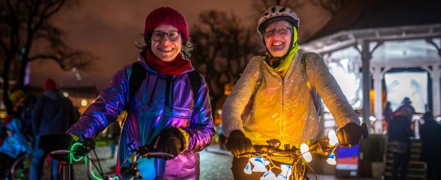 Two women stand smiling at the camera on a dark street with bikes lit up with fairy lights