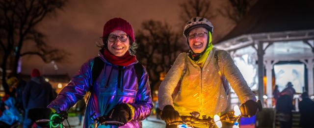 Two women stand smiling at the camera on a dark street with bikes lit up with fairy lights