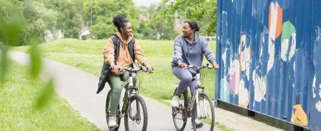 Two women cycling on a path in a park with a shipping container on their left