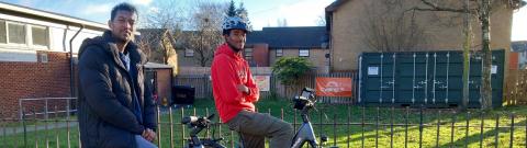 Two people sit on bicycles on a sunlit residential street, paused beside a grassy verge with low brick buildings and a fenced utility area in the background.