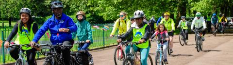 A group of adults ride bikes together through a tree-lined park in Wolverhampton during a Big Bike Revival event. Many wear high-visibility vests and helmets, smiling as they cycle in the sunshine.
