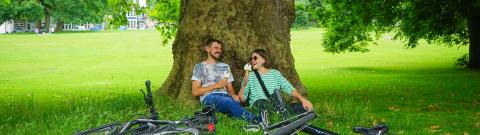 Two people sat by a tree eating ice cream, their bikes laid on the grass in front of them