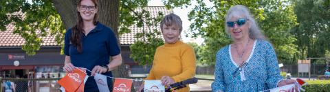 Three smiling women stand outdoors beside a yellow mountain bike decorated with Cycling UK-branded bunting. One wears a navy polo shirt, one a mustard yellow jumper, and one a turquoise patterned tunic with sunglasses. They are under a large tree with a red-roofed building in the background on a sunny day.