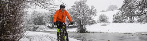Man in a bright orange jacket is cycling on a mountain bike along a snowy track