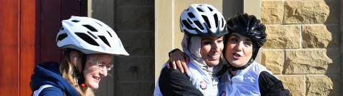 Three women in cycling helmets and white vests share a joyful moment outside a stone building. Two of them hug and smile while holding a phone, the third stands nearby with a bike, also smiling.