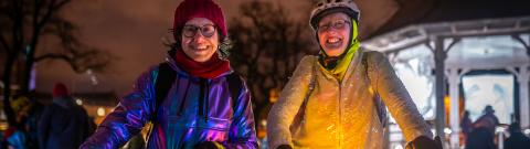 Two women stand smiling at the camera on a dark street with bikes lit up with fairy lights