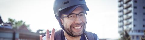 A smiling man wearing a helmet, glasses, and a cycling vest gives an ‘OK’ hand gesture while out on a sunny city street, with buildings and traffic in the background.