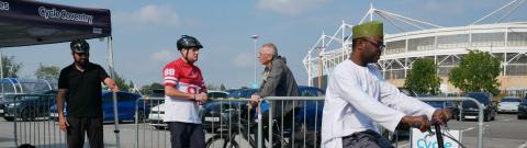 A man wearing traditional clothing and a green cap rides a small folding bike through a course marked with coloured cones in a Cycle Skills Zone in a car park. Three other men wearing helmets and casual clothing stand nearby behind a metal barrier, observing. A Cycle Coventry tent and a stadium are visible in the background.