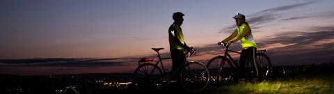 Two people stand with bikes on a hillside at dusk, almost silhouetted against the pink and grey sky