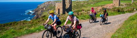 Cyclists riding along a coastal gravel trail. There are ruins and the sea in the background.