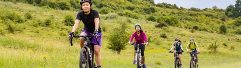Four people on touring bikes are cycling on a grassy track in the countryside. They are wearing summery clothes and cycle helmets
