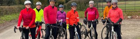 A group of people holding bikes is standing by the side of a quiet tree-lined road. They are all wearing brightly coloured cycling kit