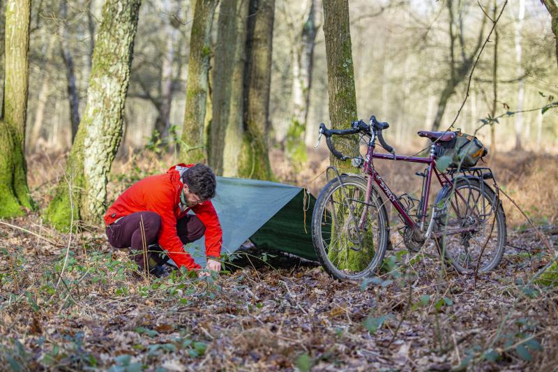 A man in a red jacket and maroon trousers is setting up a tarpaulin next to a tree in a wood to spend the night. A maroon touring bike is leaning against the tree