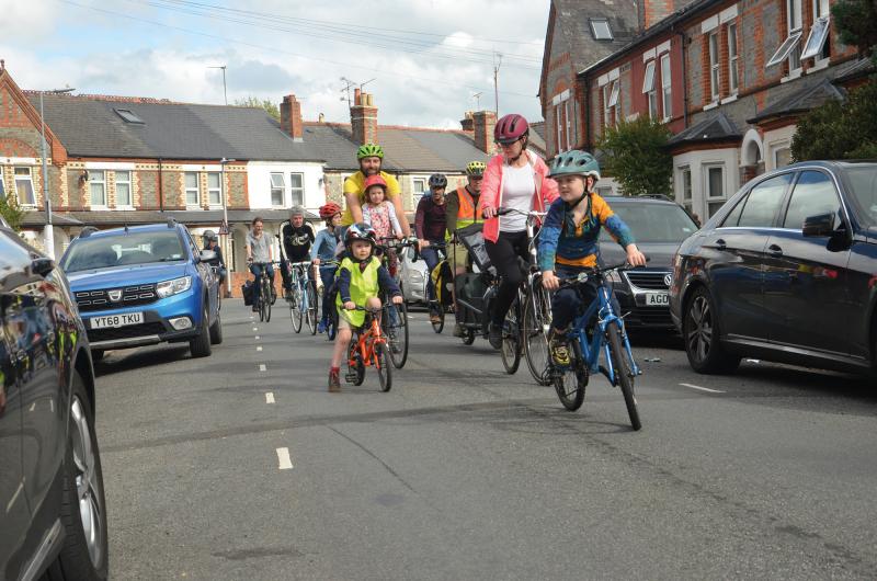 Adults and children cycling together on the road at a Kidical Mass cycling event with parked cars on wither side of the road