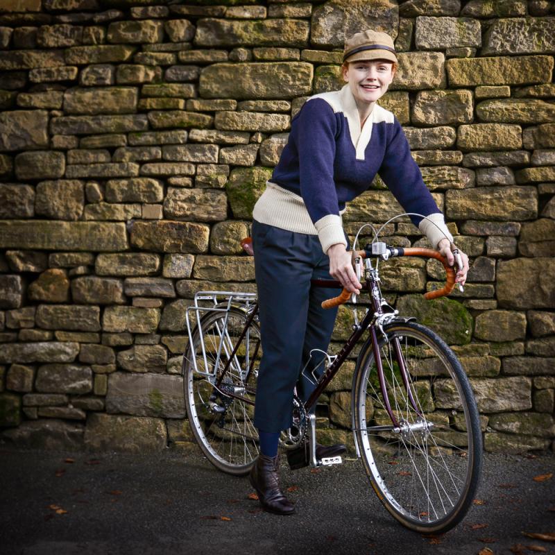 A woman is standing astride a road bike with drop handlebar and rear rack. She is wearing a flat cap, blue and white sailor top and blue trousers