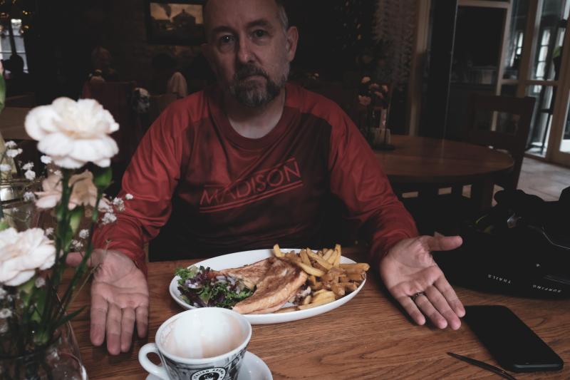 A man is sitting at a table with a plate of food in front of him. He is wearing a long-sleeved jersey. There's also an empty teacup in front of him.