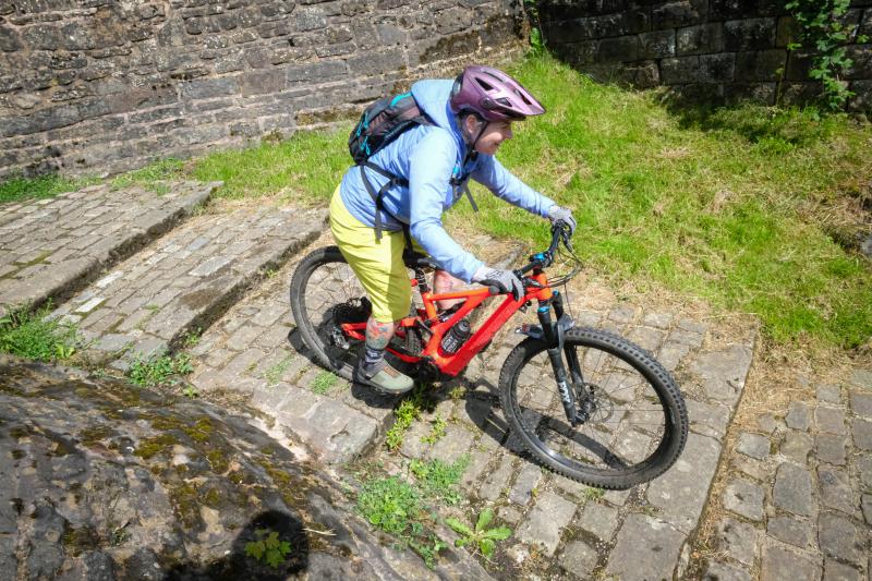 A woman on an e-mountain bike is cycling down a cobbled staircase. She is wearing waterproof cycling kit and a helmet. She has a rucksack on her back.