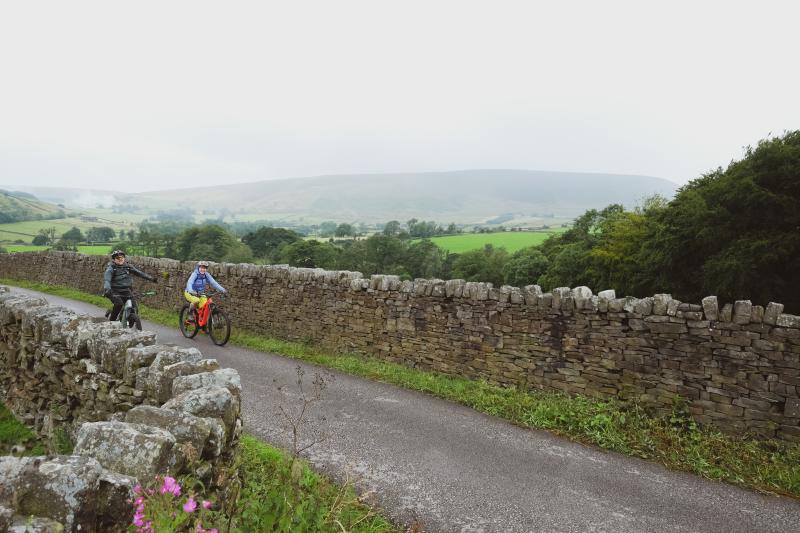Two people on e-bikes are cycling along a country lane with dry stone walls along both sides. Pendle Hill can be seen in the background