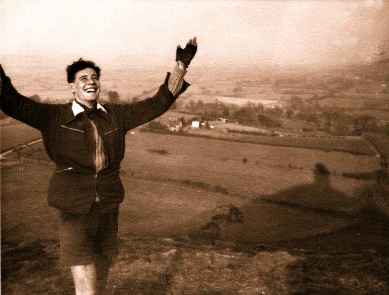 A sepia shot of a young man is standing at the top of a hill. He has his arms up and is smiling in a celebratory way. Fields and trees are laid out below him.