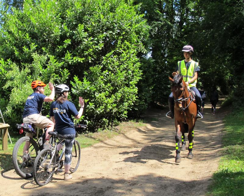 Cyclists pause on a trail to let a horse rider pass, and they exchange friendly thank you waves