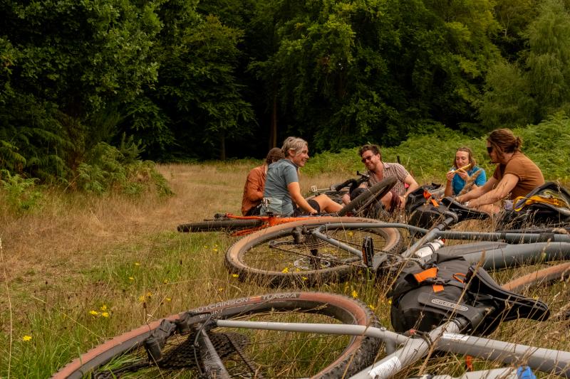 A group of people sit on the ground in a forest clearing, with bikes laid on the ground in the foreground