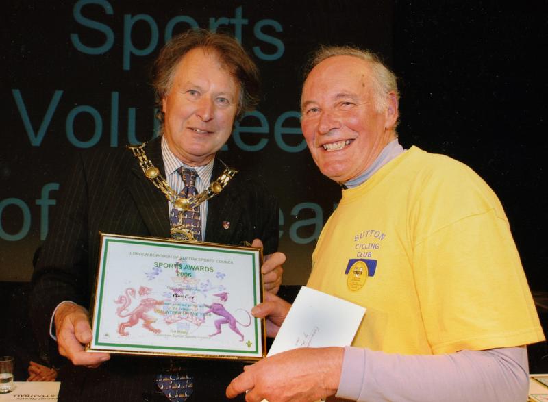 Two men pose with an award in a frame. One is wearing a suit and a chain of office. The other, receiving the award, is wearing a bright yellow Sutton Cycling Club T-shirt