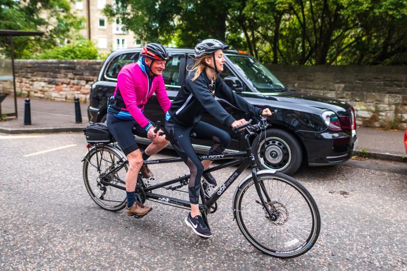 Two people are riding a black tandem on an urban street with a black cab parked in the background. An older man is on the back, a younger woman on the front. They are wearing cycling clothes and helmets.