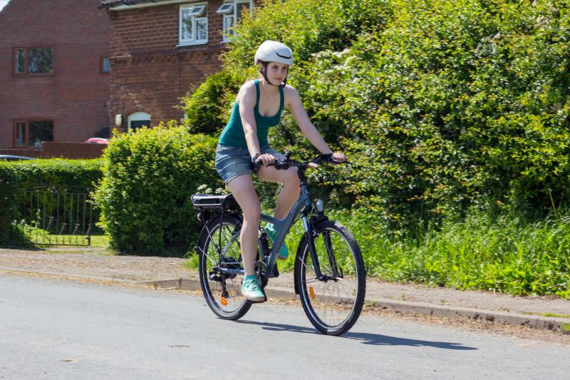 A woman is riding a grey e-bike with a flat handlebar and battery on the rear rack, in an urban setting. She is wearing denim shorts and a green strappy top and a helmet. There are houses and hedges in the background.