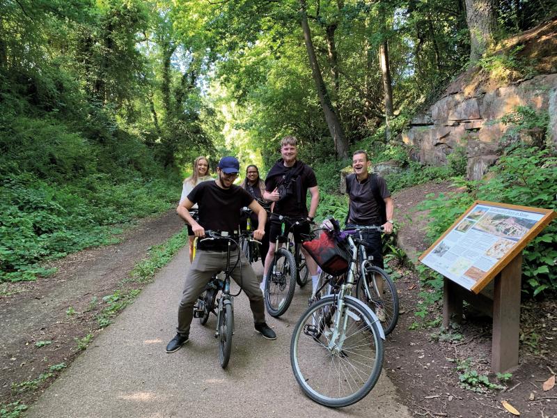 A mixed group of cyclists on a variety of bikes are on a paved path through a woodland