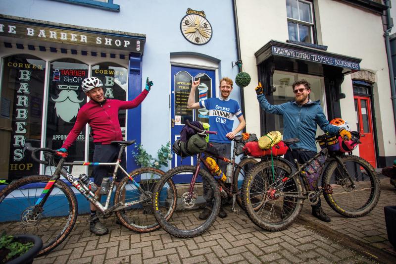 Three men and their very muddy bikes are standing in front of a barber's shop pointing up an old sign