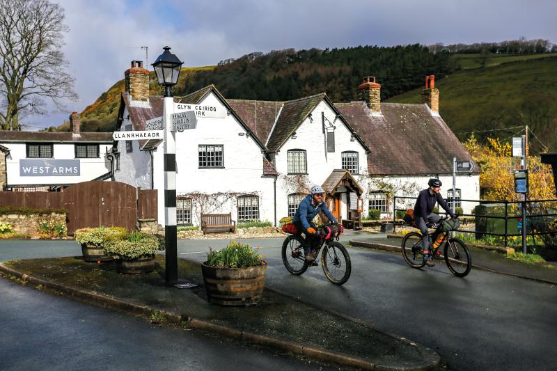 Two men are riding on a road in Wales, away from a pub