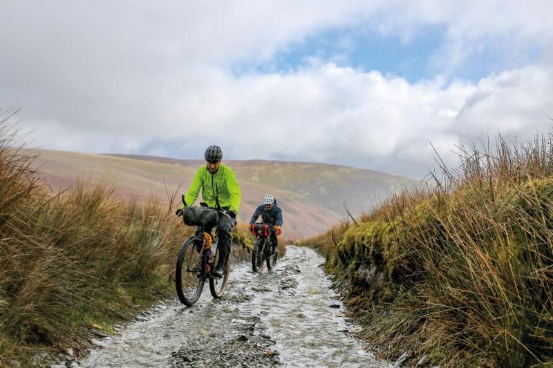 Two man are riding up a gravel track in the rain. It's very wet, there's water running down the track. They're riding packed mountain bikes and are wearing waterproofs