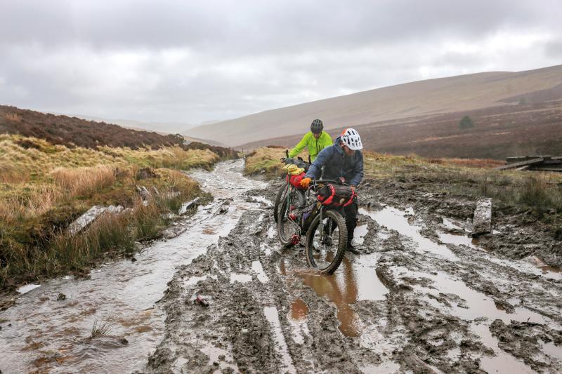 Two man are pushing their mountain bikes up a very wet, muddy trail
