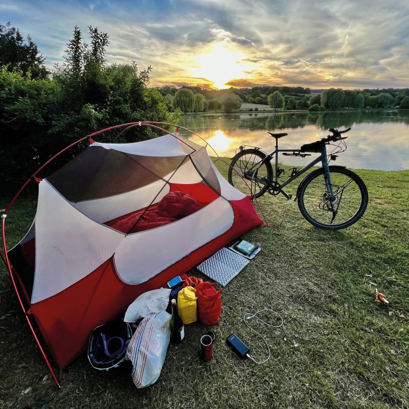 A red and white tent is pitched in front of a lake. A bike is propped up in front and camping gear is laid out beside it. It's sunset.