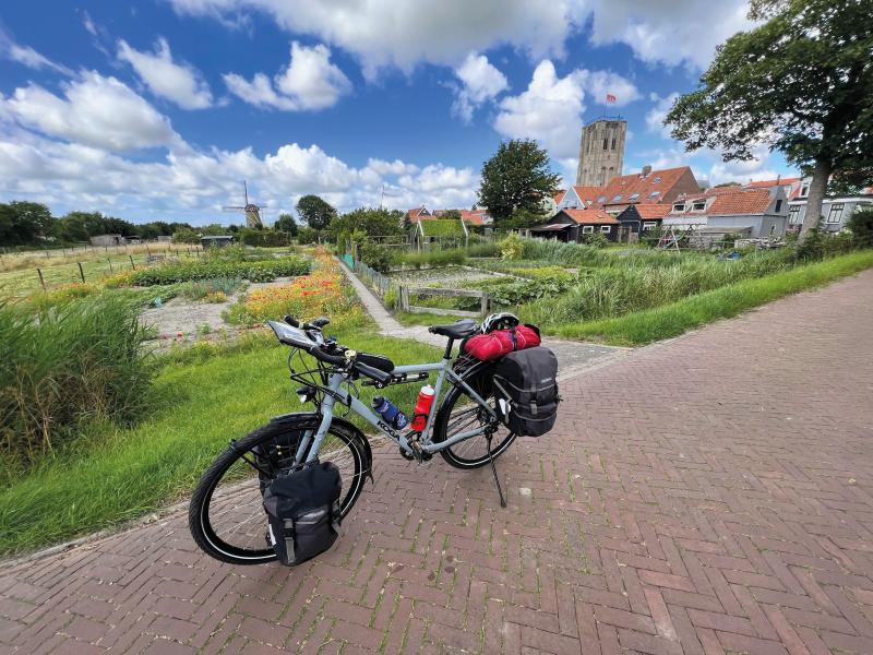 A loaded flatbar touring bike propped up on a tiled path in front of allotments with a village, windmill and church in the background