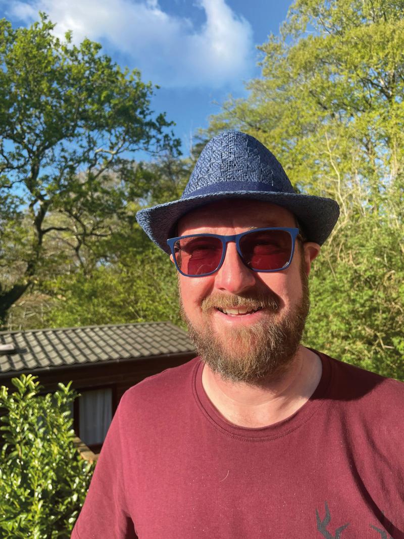 A headshot of Dave Powell. He is wearing a blue hat, sunglasses and red T-shirt. He's smiling at the camera