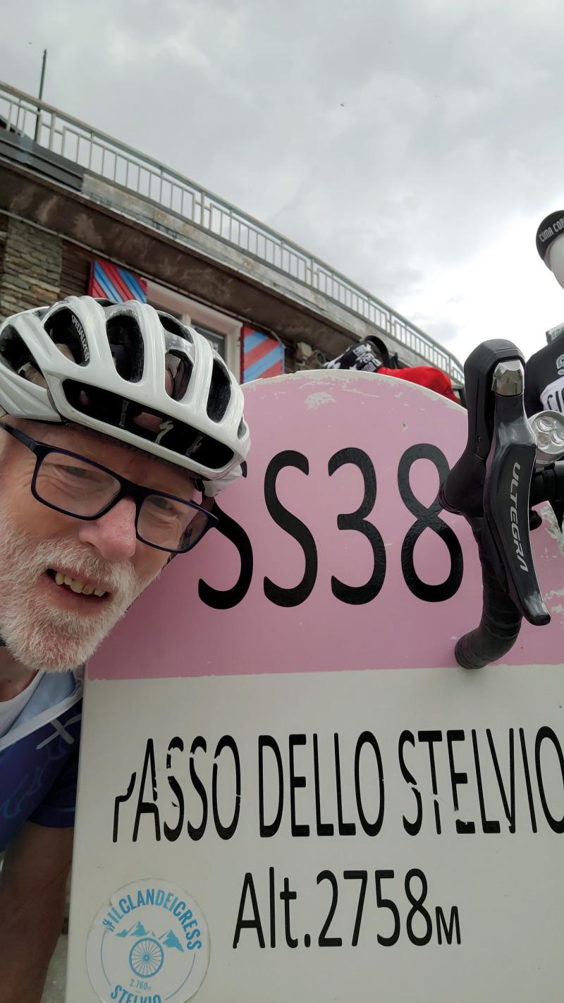 A man with glasses, a white cycling helmet and white beard is in front of a sign reading 'Passo Dello Stelvio. Alt 2758 m'. The handlebar of his bike can just be seen