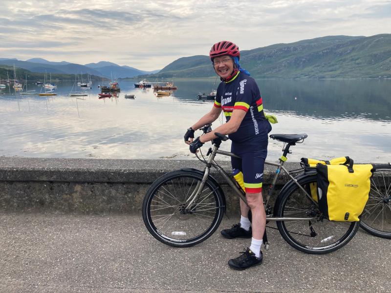 A man in full cycling kit and helmet is standing astride a flatbar touring bike with packed panniers. He is on a concrete bridge across a waterbody. There are mountains in the background.