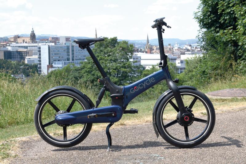 A blue e-folding bike propped up on a gravel path with an urban environment in the background