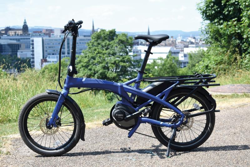 A blue e-folding bike propped up on a gravel path with an urban environment in the background