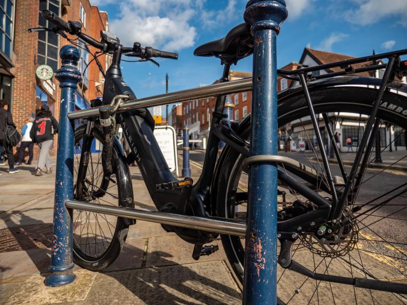 A black Raleigh e-bike is locked to a cycle hoop in a high street. There is one D-lock through the back wheel and one around the frame