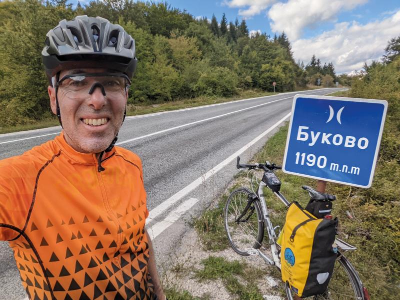 A man in an orange cycling jersey with black triangle pattern is taking a selfie showing his loaded touring bike and a road sign. He is wearing a cycle helmet and glasses