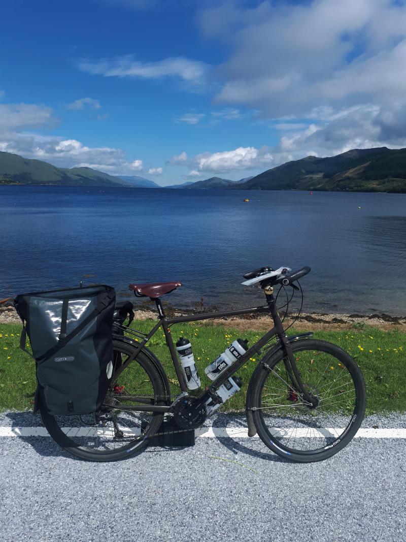 An Oxford Bike Works tourer is upright on a road in front of a large lake surrounded by mountains