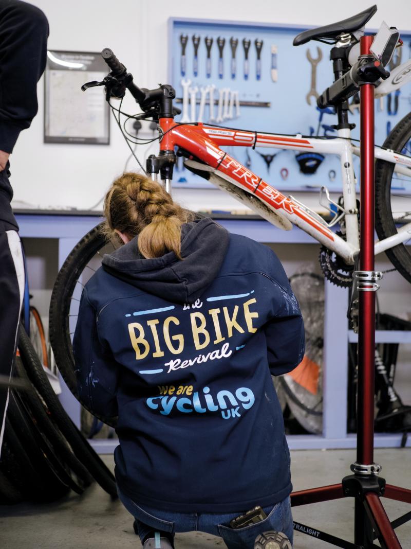 A woman is working on the front wheel of a red and white bike that is raised on a bike stand. On the back of her jacket is the Big Bike Revival logo