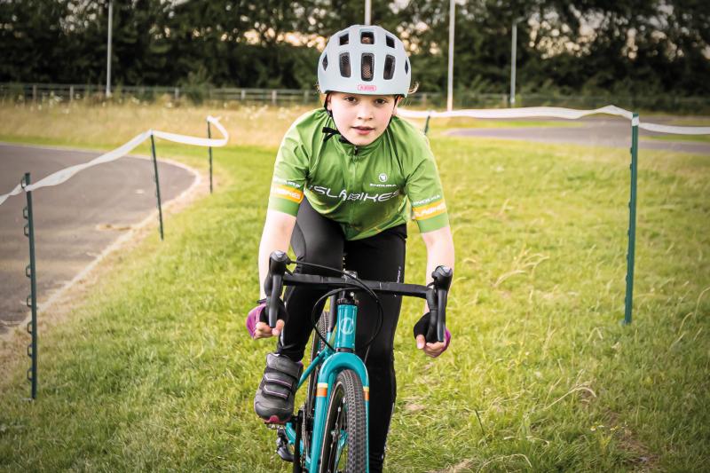 A young girl is riding an Islabike, wearing a green cycling jersey and grey helmet