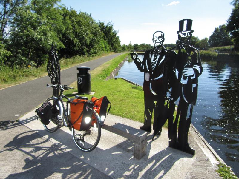 A loaded flatbar touring bike with two very large panniers is leaning against a bench next to a metal silhouette of two men, one of whom is wearing a top hat