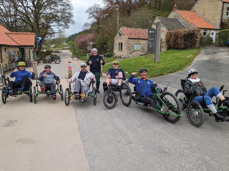 A group of people are all on non-standard cycles, including trikes, recumbents, quadricycles. They are in a country road in a village.