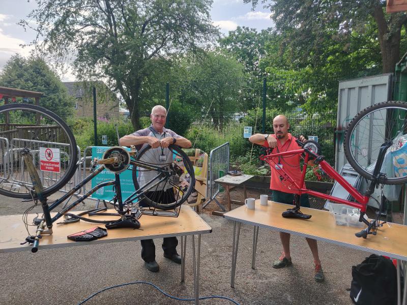 Two men are fixing bikes. The bikes are upside down on trestle tables and one man is refitting an innertube, the other is adjusting the chain. There are two mugs of tea on the tables.