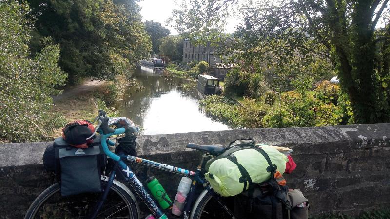 A loaded Dawes Galaxy touring bike is leaning against a stone wall of a bridge going over a canal, which is lined with trees and bushes